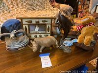 Overview of assortment of animal art pieces on wooden table including dolphin sculpture, wooden rhino, stuffed moose toy, bird sculptures, bear head art, and decorative drawer set