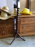 Three new straw hats placed on a wooden cabinet: two natural straw hats and one yellow safari hat. Center of image shows part of the dark wooden hat tree column with twisted design.