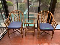 Set of two rattan armchairs with blue cushions and one matching rattan end table with glass top arranged side by side indoors on terracotta tiles.