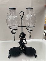 Front view of the double glass wine decanter set on black metal stand with leaf decorations and two concave trays at base.