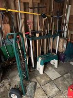 Photo showing variety of gardening tools including shovels, rakes, and garden fork, two hand trucks/dollies, and a green plastic step stool.