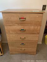 Front view of wood highboy dresser with five drawers, top drawer handle has red accent, lower drawers have green and blue accents on brass handles.