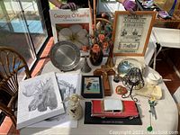 Wide view of all items in the lot as shown on table including books, framed art, figurines, model car, lampshade, and drawings.