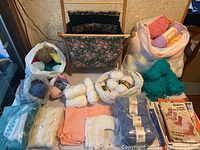 Wide view of multiple yarn skeins and balls in various colors including white, teal, and pink with knitting patterns and fabric pieces scattered.