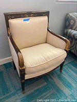 Full view of beige upholstered armchair with dark wood frame and gold decorative carvings placed on carpeted floor.