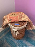 Round wicker laundry basket with fabric lining and patterned cloth draped over the lid in a corner with blue carpet and purple walls.