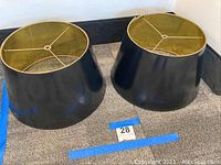 Two black lamp shades with gold interiors, placed on carpeted floor with damage visible.