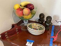 Glass fruit bowl with faux fruit, two metal busts, and three stacked decorative plates on wooden surface