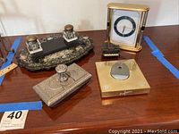 Lot items arranged on table: ink well set with two glass inkwells on black ornate blotter, silver-tone engraved ink well, gold-tone Wako clock, piano pencil sharpener, and brass pen holder.