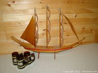 Wooden sailboat model with three masts, sails, and mounted stand next to three brown Siesta Ware mugs with white nautical ship design on a wooden backdrop.