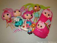 Wide shot showing four dolls standing and two pink rolled blankets with Lalaloopsy printed images, plus a small orange and black toy figure.