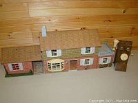 Front view showing the two-story dollhouse with a bay window, chimney base (chimney missing), and attached smaller structure with brown roof.