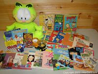 Full view of Garfield plush toy along with an assortment of comic books and books featuring Garfield and other comic strips, laid out on a wooden surface.
