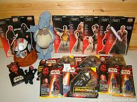 Wide photo showing multiple Star Wars Episode 1 items including standees of Darth Maul, Sith, Watto, Jar Jar, several action figures in original packaging, a plush toy, and a helmet on a wooden stand.