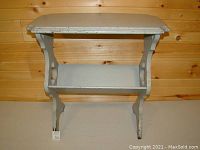 Full view of small wooden end table with curved sides and lower shelf. Pale gray distressed paint with wear visible.