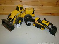 Photo showing two Tonka branded yellow metal construction vehicles on a white surface against a wooden wall background. Vehicles include a front loader with large rubber tires and a tracked excavator.