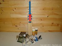 Full view of cricket bat with blue grip, tubes of birdies, bag of golf tees, and bike lock on white surface with wood wall background.