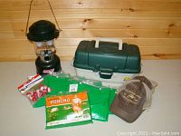 Photo showing the complete lot with Coleman lantern, Plano tackle box, fishing bobbers, two hats, and several new green ponchos in packaging.