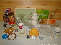 Wide view showing mixed lot of glass, ceramic, and metal items arranged on table against wood wall