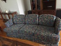 Photo of fabric upholstered couch with floral, vine pattern in blue, red, beige, and green, featuring three back cushions and rolled arms.