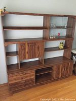 Front view of large wooden particle board cabinet with multiple shelves, two cupboards, and glass doors. Several small decorative items and glassware placed on shelves.