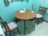 Wood-grain round table with two wooden chairs having padded cushions, placed in a room with teal walls and tiled floor