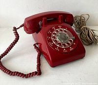 Red rotary dial telephone on white surface showing front view with handset, dial, and cords visible.