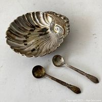 Photo of an antique scallop shell shaped sterling silver salt cellar with two matching small spoons on a light surface, showing details and tarnish.