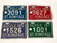 Four vintage metal bicycle license plates from St. Boniface dated 1967, 1969, 1970, and 1971 arranged side by side on a white backdrop.