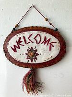 Full view of the vintage oval welcome sign hanging on a wall, showing carved "WELCOME" text, central floral/star motif, hanging string with beads, and tassel.