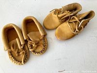 Two pairs of light tan leather child shoes, moccasins style. One pair with fringe and laces, the other with slip-on style and lace ties.