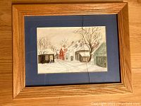 Framed watercolor painting depicting snowy village street with houses, trees, and bicycle, in wooden frame with visible crack in glass.