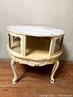Front angled view of oval cream painted vintage French Provincial style table with distressed finish. Shows open chicken wire panel side sections and curved cabriole legs.