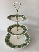 Full view of the three-tier dish from front showing the green floral boutonniere design on white plates and the gold-tone stand with triangle handle on top.