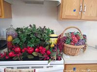 Photo showing faux floral garland with red berries and pomegranate-like faux fruit on a kitchen countertop next to a yellow biscuit jar and basket of faux grapes.