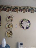 Ceramic decorative plates with fruit and grape design vertically arranged on a white metal rack against a white wall under a decorative grape and leaf wallpaper border.