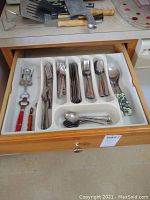 Drawer open showing stainless steel silverware organized in white plastic tray with knives, forks, spoons, and serving utensils. Adjacent kitchen utensils and bottle openers visible on countertop.
