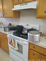 Wide kitchen shot showing several pots and pans on a stove and counter, including a large aluminum pot and multiple dark green pots with lids.