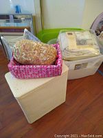 Laundry basket with pink floral liner, decorative cushion on padded storage cube, packaged bedding beside basket