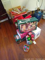 Photo of stacked boxes of Christmas decorations on floor, includes Bayberry Village box, glass ornaments in bowl, lamp in background
