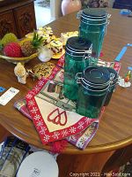 Four green glass canisters of different sizes on top of a folded, patterned table runner with decorative items around.