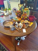 Wide view of the wooden table displaying ceramic pots with fruit relief, ceramic beer steins, copper pan and measuring cup, and other ceramic items.