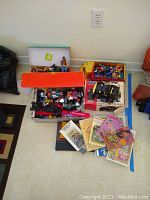 Large collection of vintage toys and books displayed on floor including boxes filled with action figures, toy trucks, and scattered books.