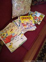 Collection of vintage magazines and comic books on a red couch, includes Mad Magazines, Cracked comic, and others.