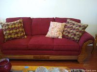 Front view of three seat burgundy velour sofa with three attached decorative pillows and carved wood framing along bottom and arms.
