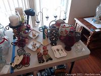 Image showing an assortment of decorative items including mosaic candle holders, two figurines, ceramic plates, and metal candle holders on a white folding table.