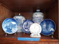 Display of four Danish Christmas plates, porcelain clock, and two blue and white porcelain jars on a wooden shelf.