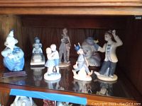 Wide shot of eight Mexican porcelain figurines displayed inside a wooden cabinet including clowns, children, and adult characters in various poses and outfits.