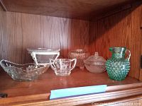 Shelf with six cut glass items including a green hobnail pitcher, a candy dish, a sugar bowl and three clear glass bowls