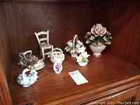 Overview of Capodimonte porcelain pieces including floral arrangements, a small chair, basket, swan and shoe-shaped figures on wooden shelf.